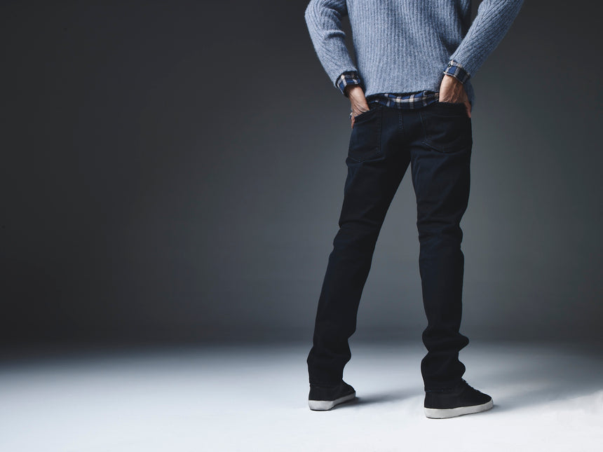 Back shot from shoulder down of a model standing with his hands in his back pocket against a dark background wearing a grey sweater and S.M.N Studio Bond in Shadow jeans. 
