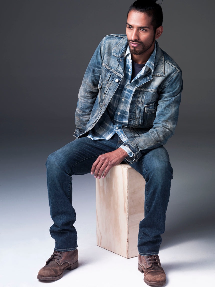 Ponytailed and bearded dark skinned model sitting on wooden box while leaning over to his left side. He is wearing S.M.N Studio's Trucker Jacket in Morrison with a plaid shirt underneath, Bond in Odyssey slim straight jeans, and dark brown boots.