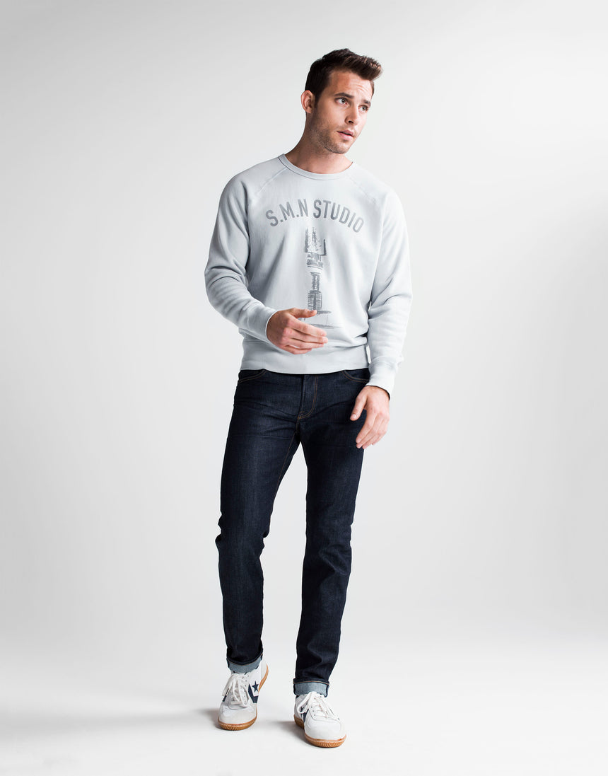 Brown haired athletic model standing wearing S.M.N Studio's Bond in Bravo jeans and S.M.N Studio printed grey crewneck sweatshirt. The jeans are a slim straight raw denim washed jean. 