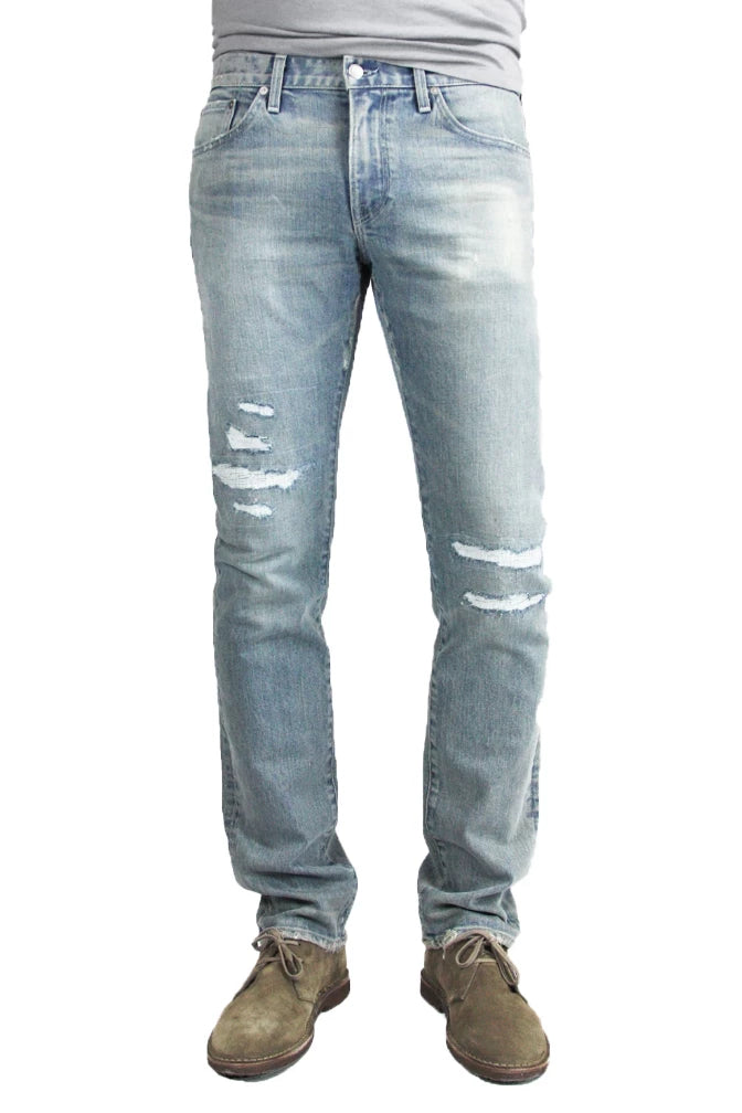 S.M.N Studio's Bond in Vernon Repair men's jeans. A slim straight jean made in a comfort stretch Japanese denim. Its light blue wash is contrasted with fading and repaired distressing around the knees with subtle rip details on the hems of the jeans.