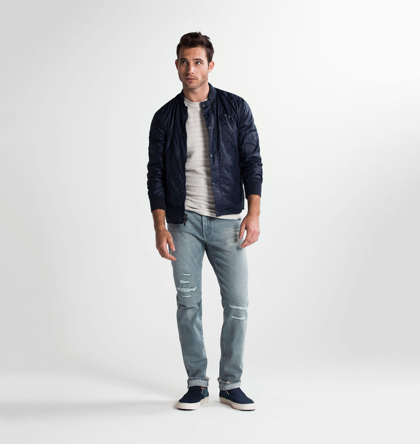 Brown haired athletic model standing and slightly bending his left knee while wearing a navy bomber jacket rolled up at his sleeves with a grey t shirt and S.M.N Studio's Bond in Vernon Repair jeans. The jeans are a slim straight jean in a light blue wash with repaired distressing at the knees and subtle rip details on the hems. 