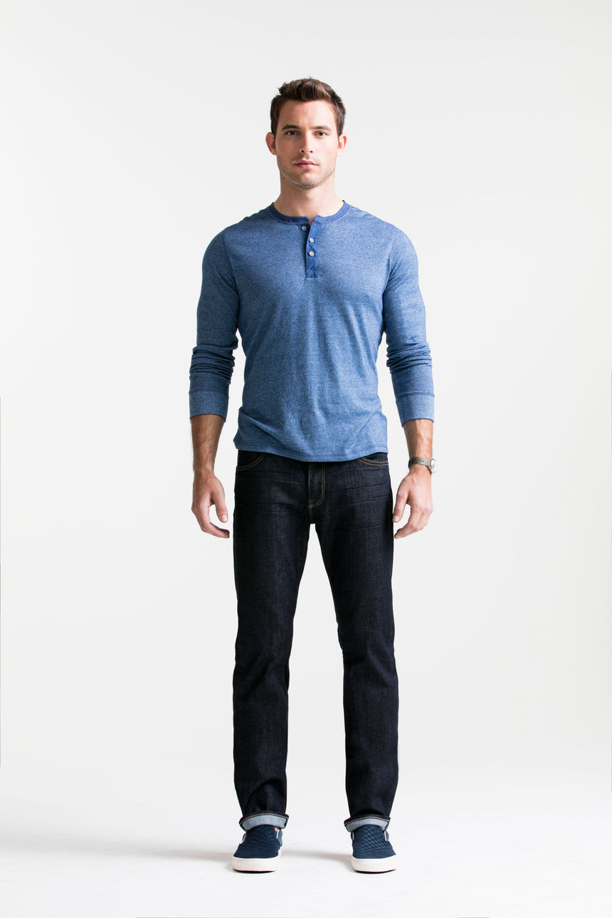 Brown haired athletic model standing in SMN Studio's Bond in Bravo Men's Jeans and blue henley. The jeans are a slim straight raw denim washed jean. 