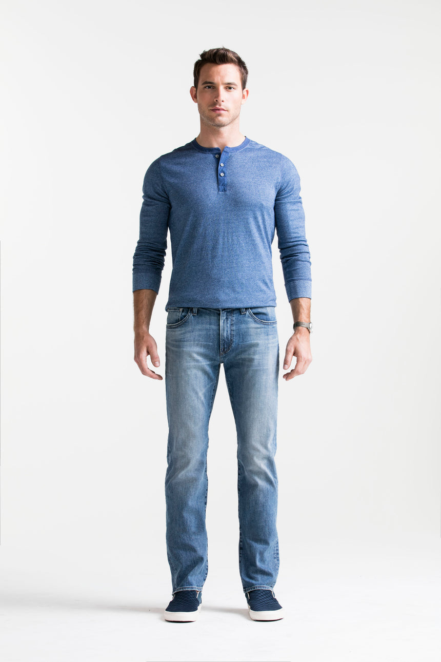 Brown haired athletic model standing in S.M.N Studio's Bond in Costello men's jeans and blue henley. The jeans are in a slim straight light blue vintage washed denim.