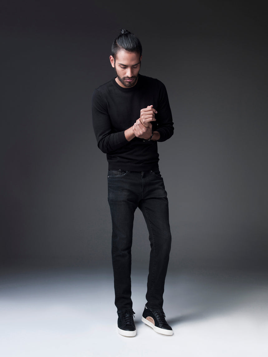 Ponytailed model dressed in all black with a black sweatshirt, S.M.N Studio's Finn in Black Rock, and black tennis shoes. He's in front of a heavily shadowed background grabbing his right wrist with his left hand.  